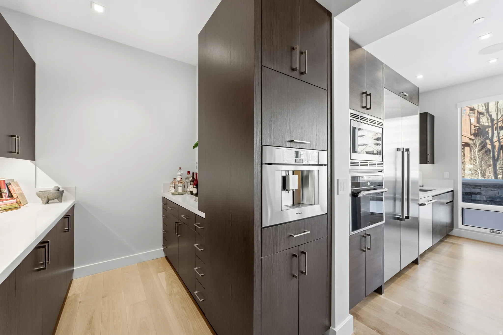 Modern kitchen with warm wood-finish flooring, dark wood cabinetry, white countertops, built-in stainless steel appliances, and an integrated coffee system