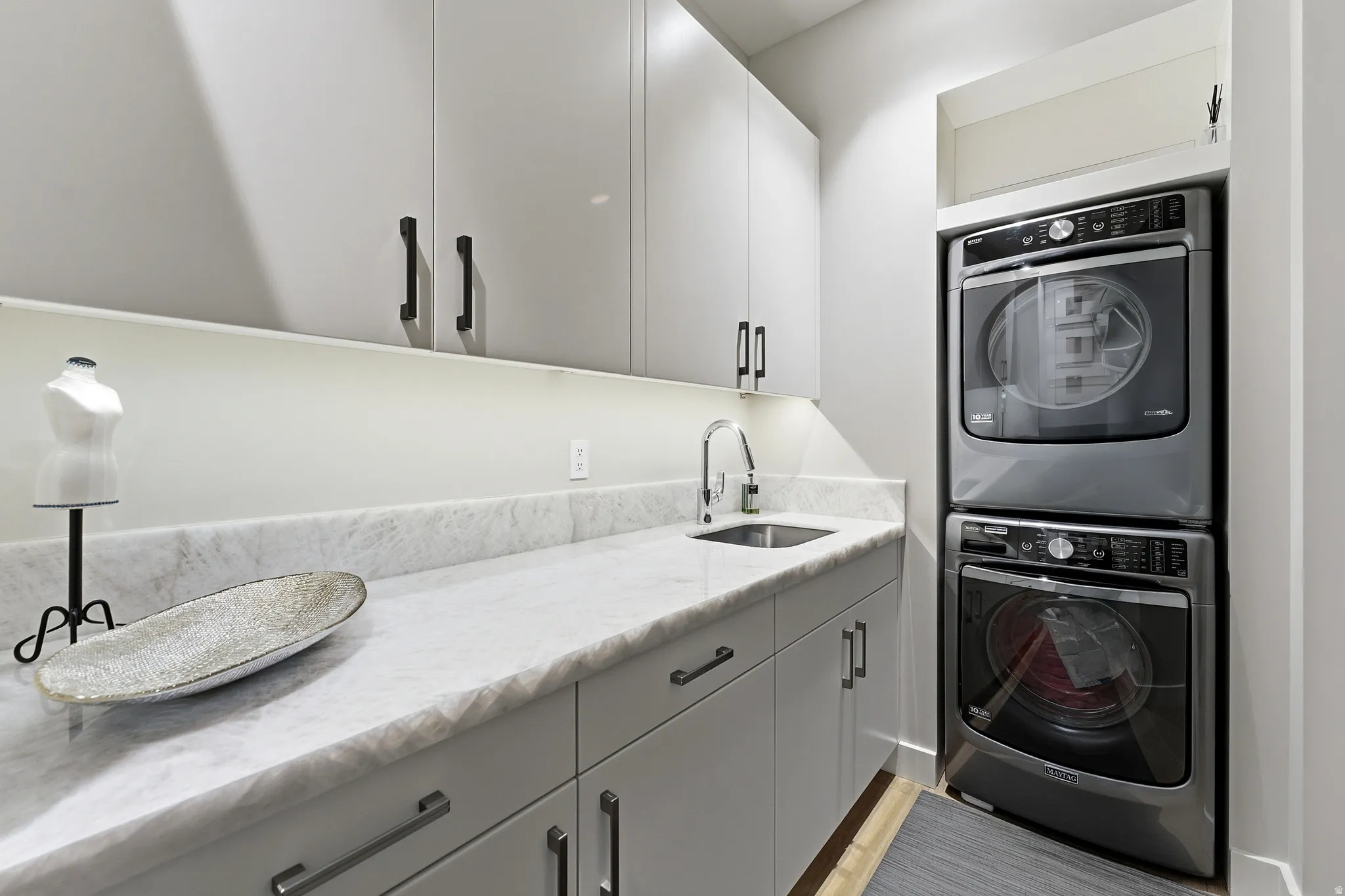Laundry room featuring a stacked washer and dryer, extensive countertop space, integrated sink with gooseneck faucet, upper and lower cabinetry, and light wood-finish flooring