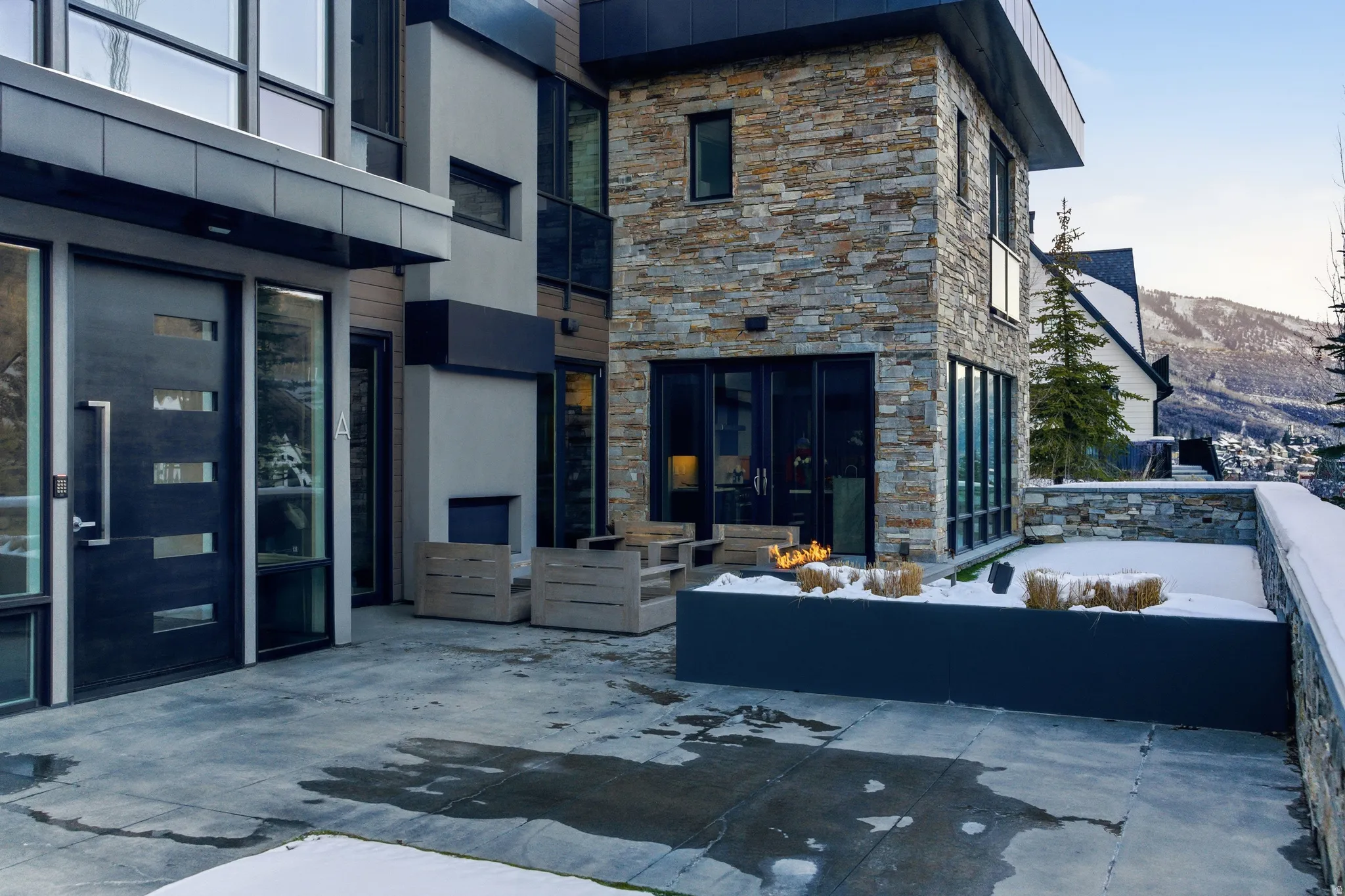 Modern architectural exterior featuring stone and stucco finishes, large glass panels, a sleek entry door with horizontal lites, and an exterior fireplace feature