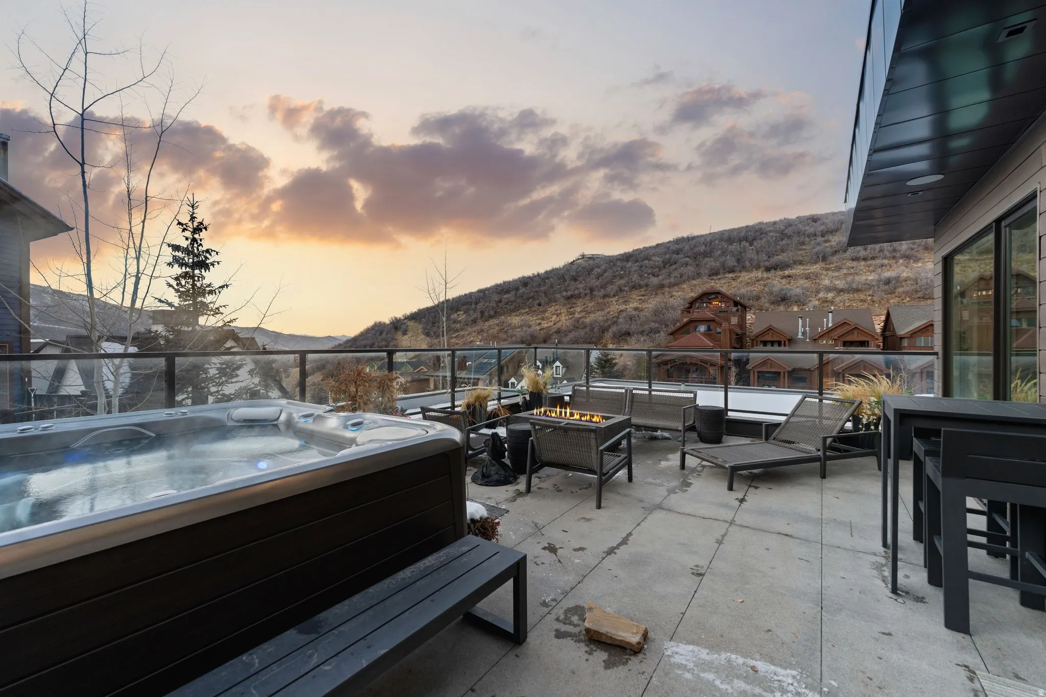 Expansive outdoor patio featuring a built-in hot tub, concrete tile flooring, glass railing, and a gas fire pit