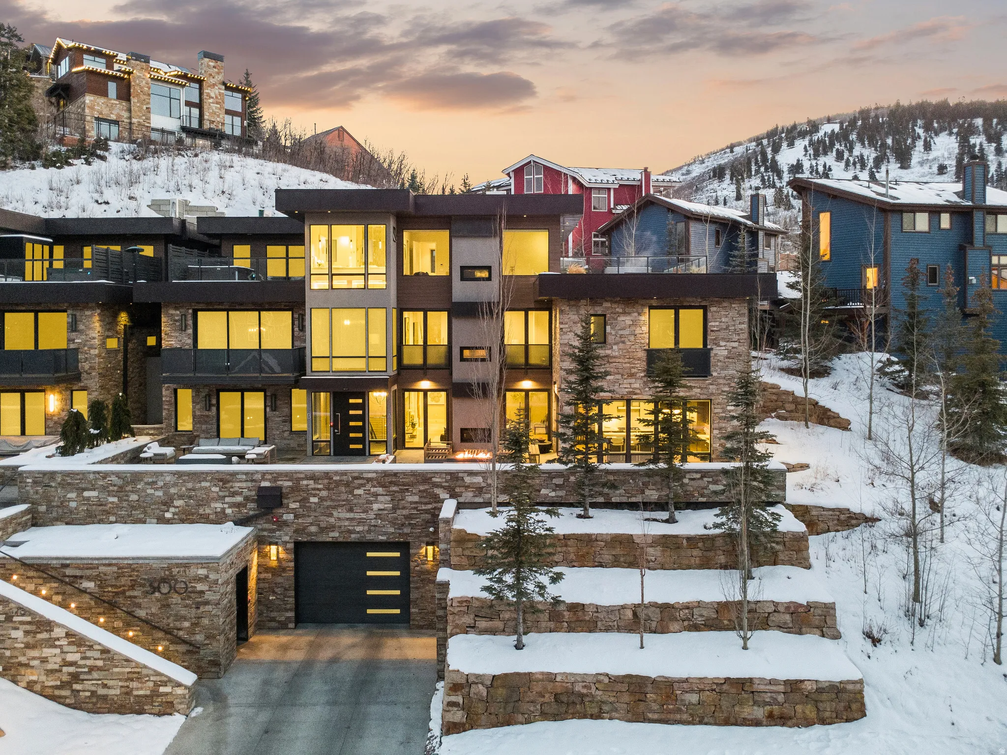 Multi-level contemporary residence featuring extensive stone veneer, expansive window walls, multiple balconies with glass railings, a recessed garage with a modern door, and illuminated exterior steps