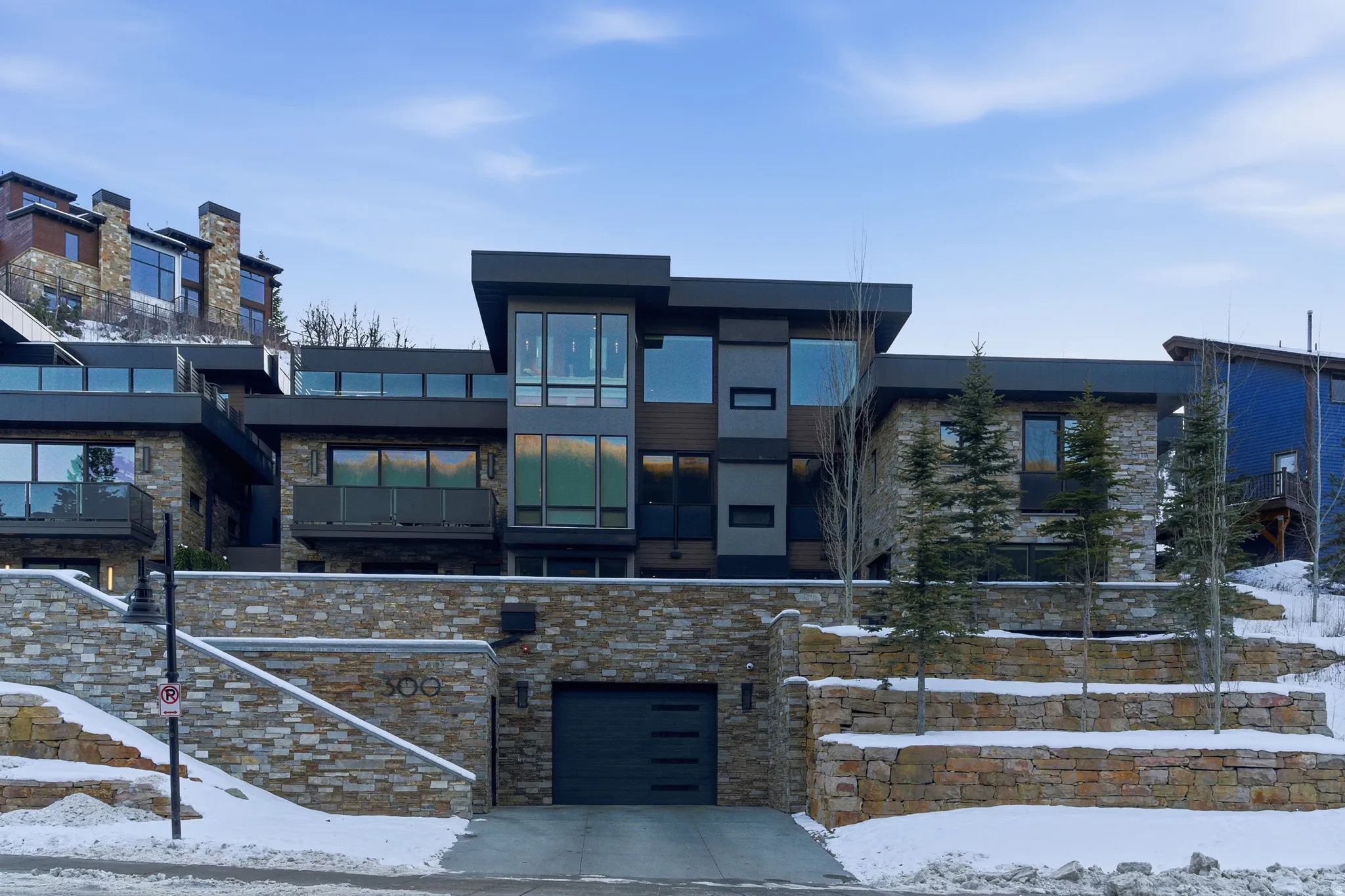 Multi-level modern residence featuring extensive stone facade, oversized windows, multiple balconies with glass railings, a built-in garage with a contemporary door, and terraced landscaping
