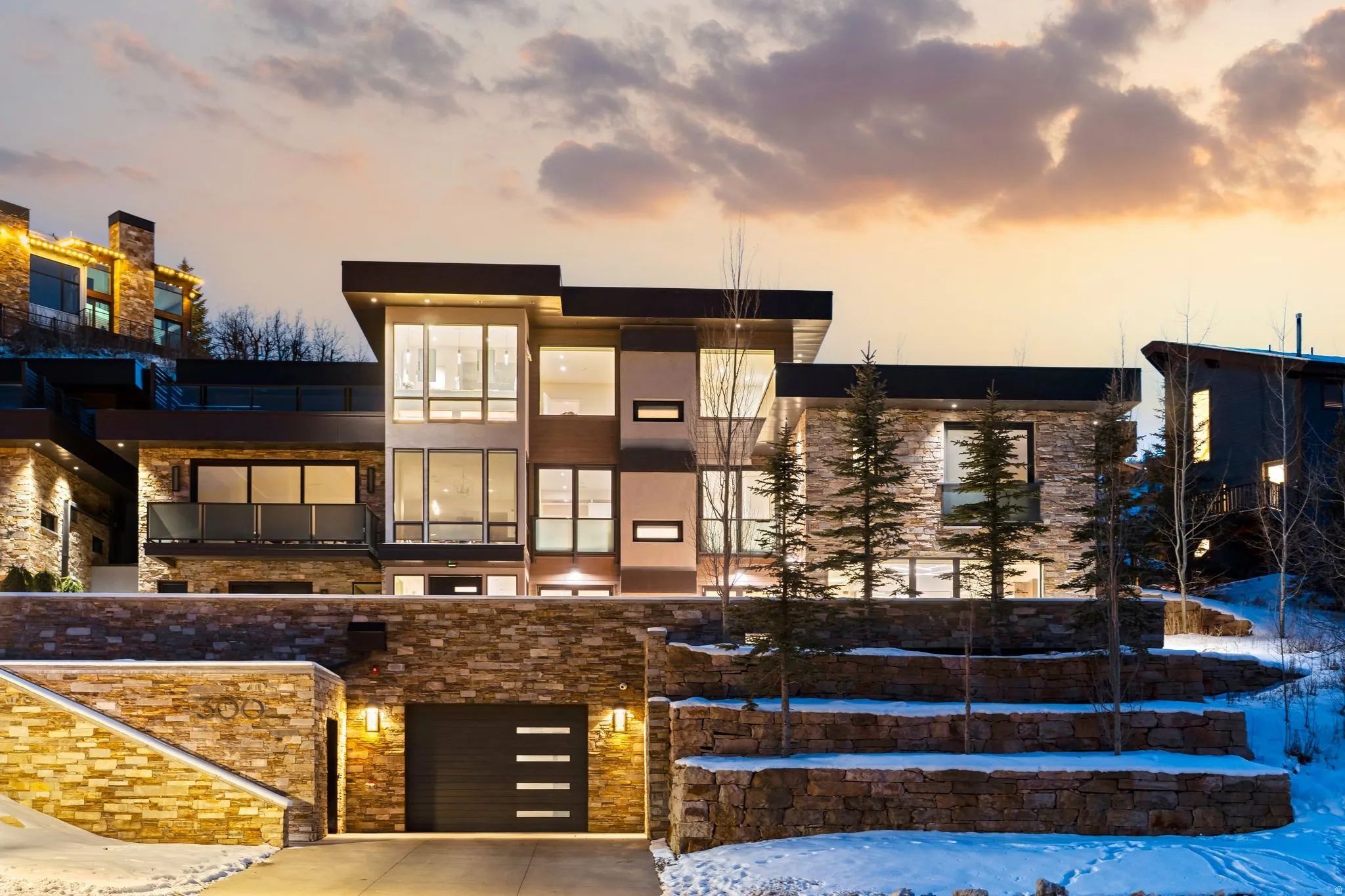 Contemporary multilevel residence featuring extensive stone veneer, a sleek black garage door with horizontal accents, multiple large windows, and an upper-level balcony with a glass railing