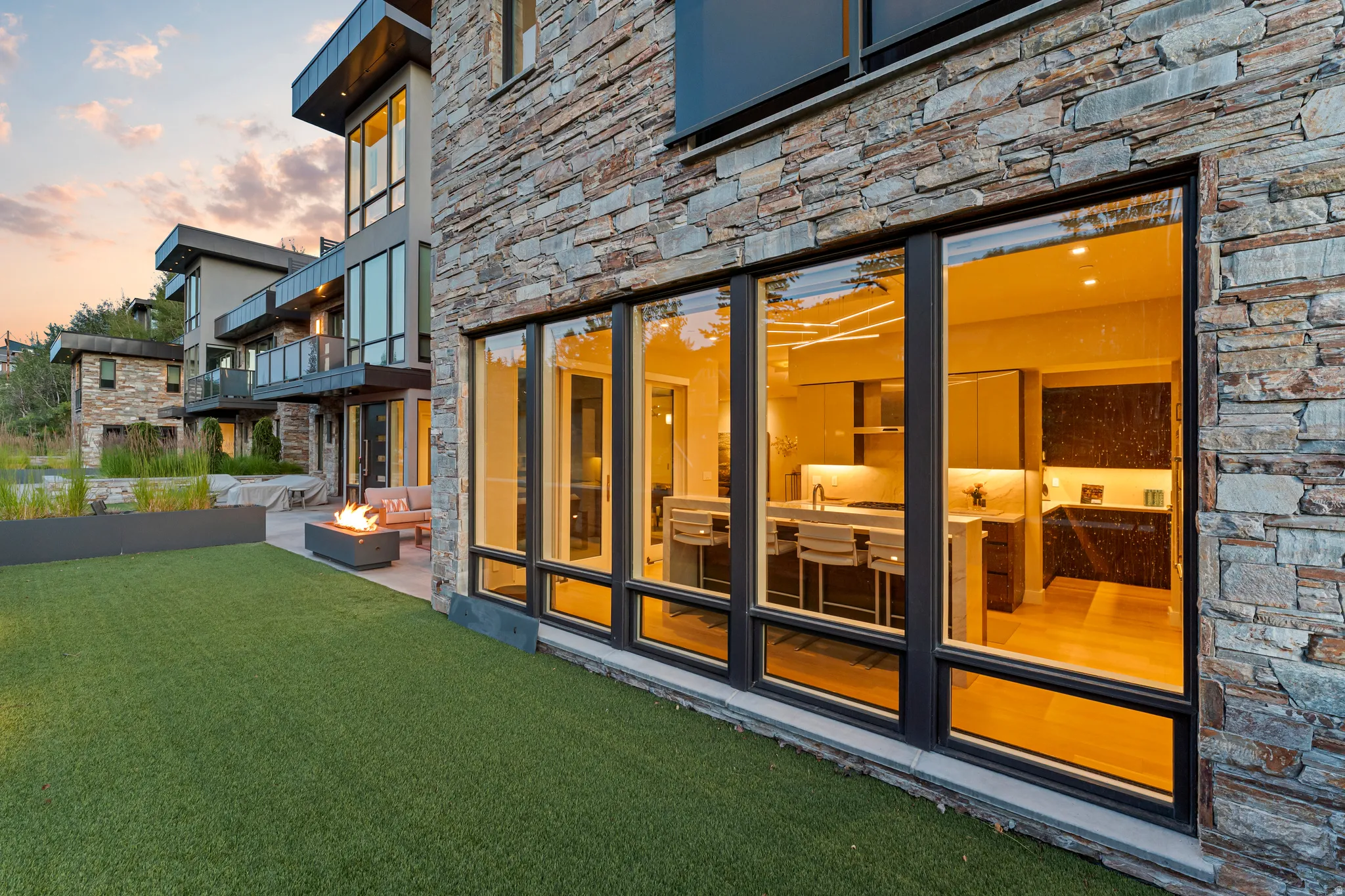 Stone veneer exterior with expansive black-framed windows revealing an illuminated kitchen interior