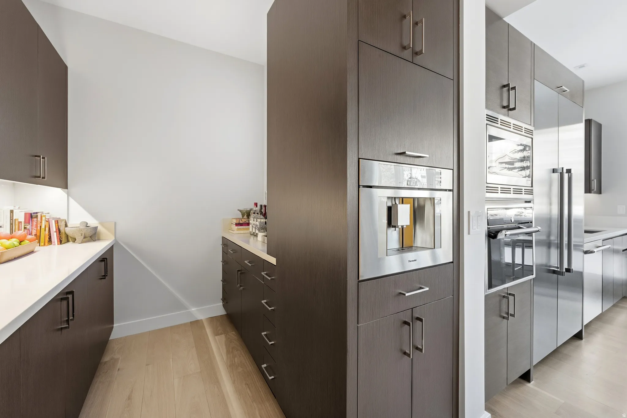 Contemporary kitchen featuring dark wood-finish cabinetry, light wood-finish flooring, white countertops, built-in stainless steel appliances, and integrated under-cabinet lighting
