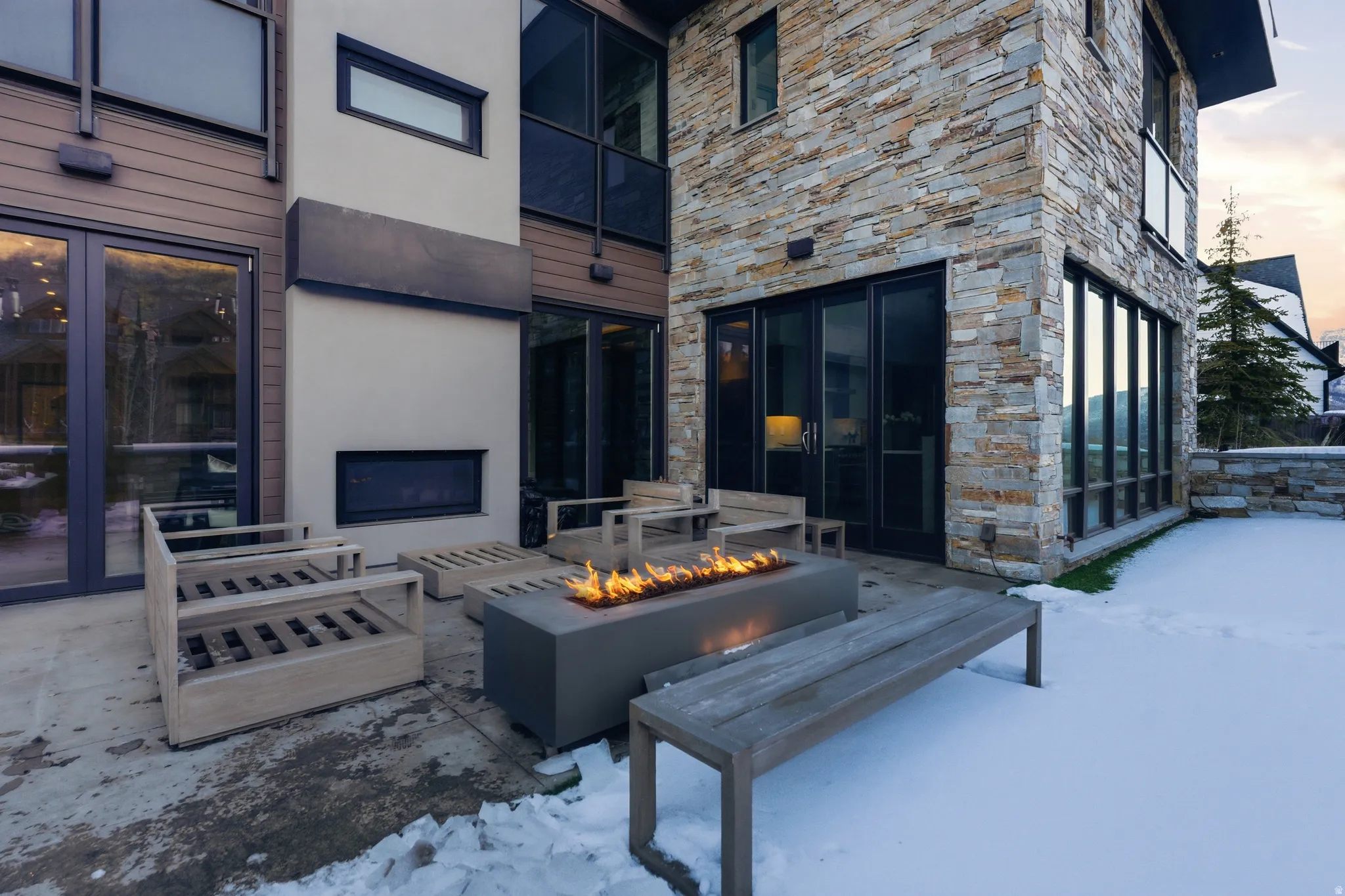 Contemporary stone and wood-paneled exterior featuring an outdoor fire pit, multiple large windows, and patio access