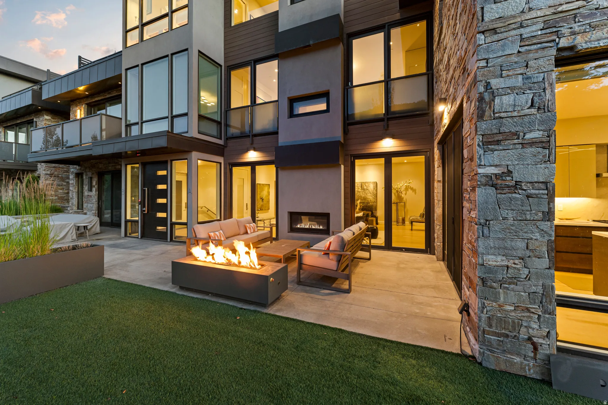 Modern exterior design featuring large format windows, a stone accent wall, a concrete patio with a built-in fire pit, a turf lawn, and a multi-level structure