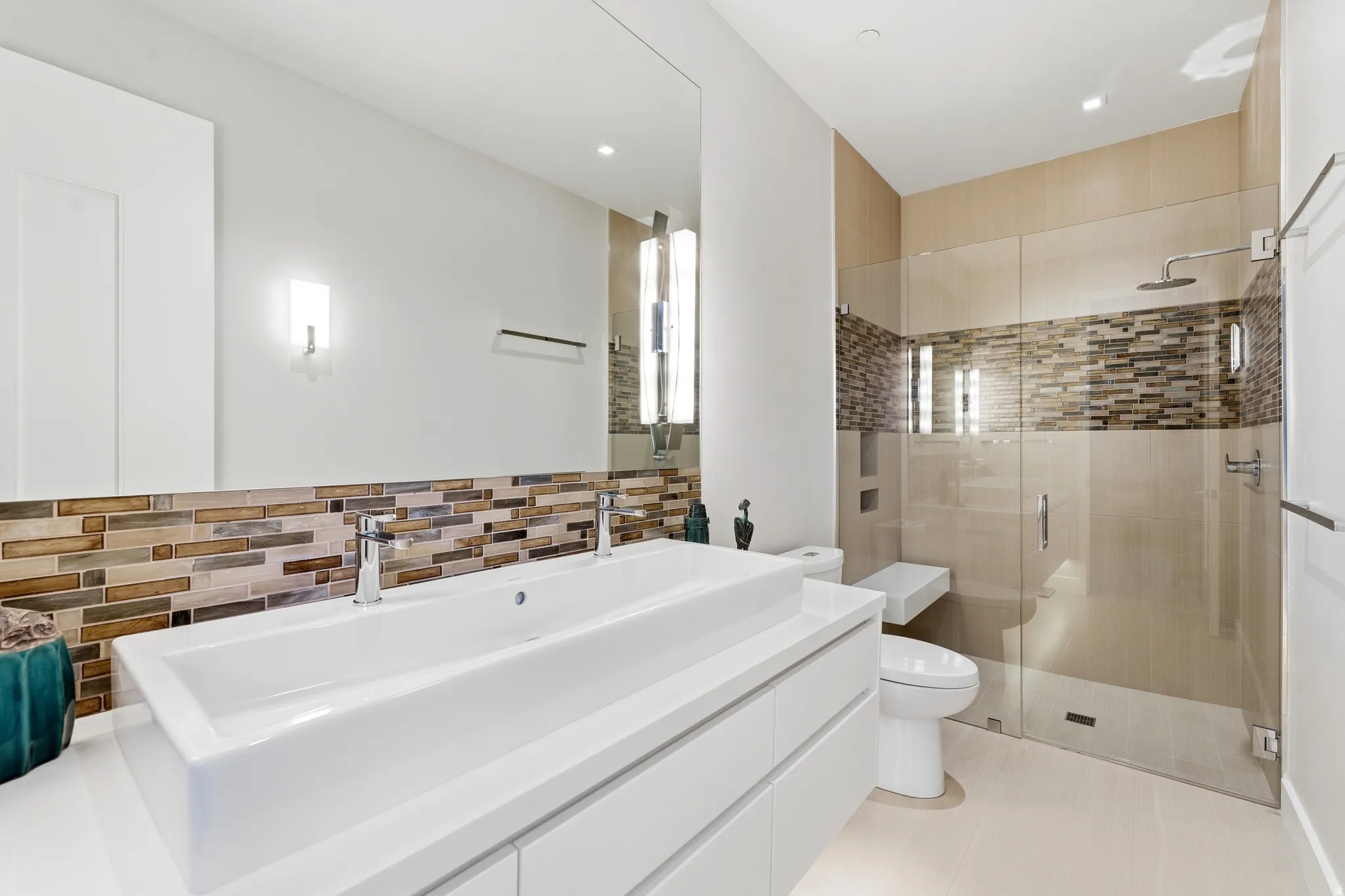 Contemporary bathroom featuring a dual vanity with rectangular vessel sinks, extensive mirrored wall, glass-enclosed shower with a built-in bench, and a mosaic tile accent band