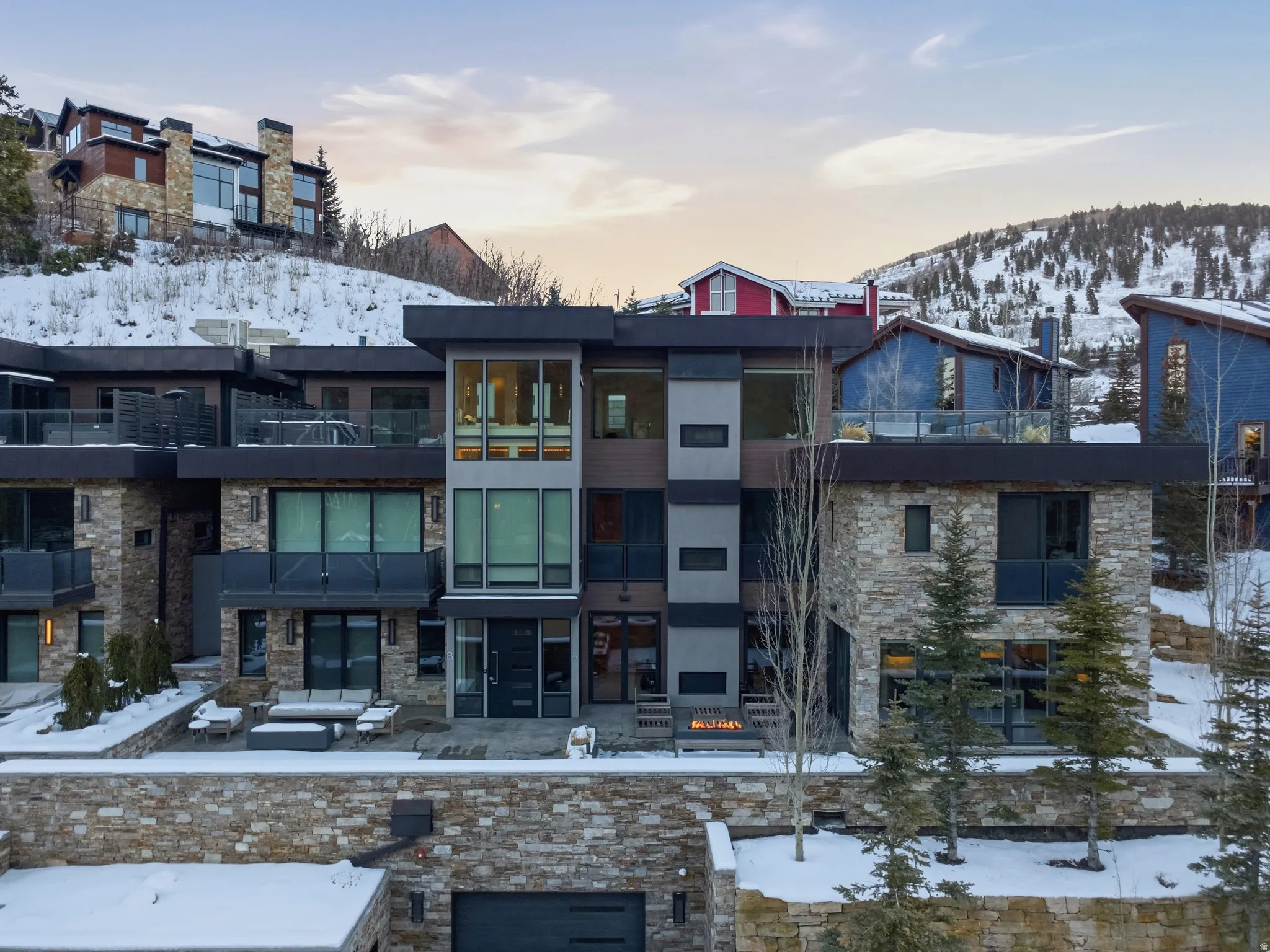 Contemporary residence featuring stone and dark panel siding, multiple balconies with glass railings, a stone retaining wall, an attached garage, and mountain terrain in the background