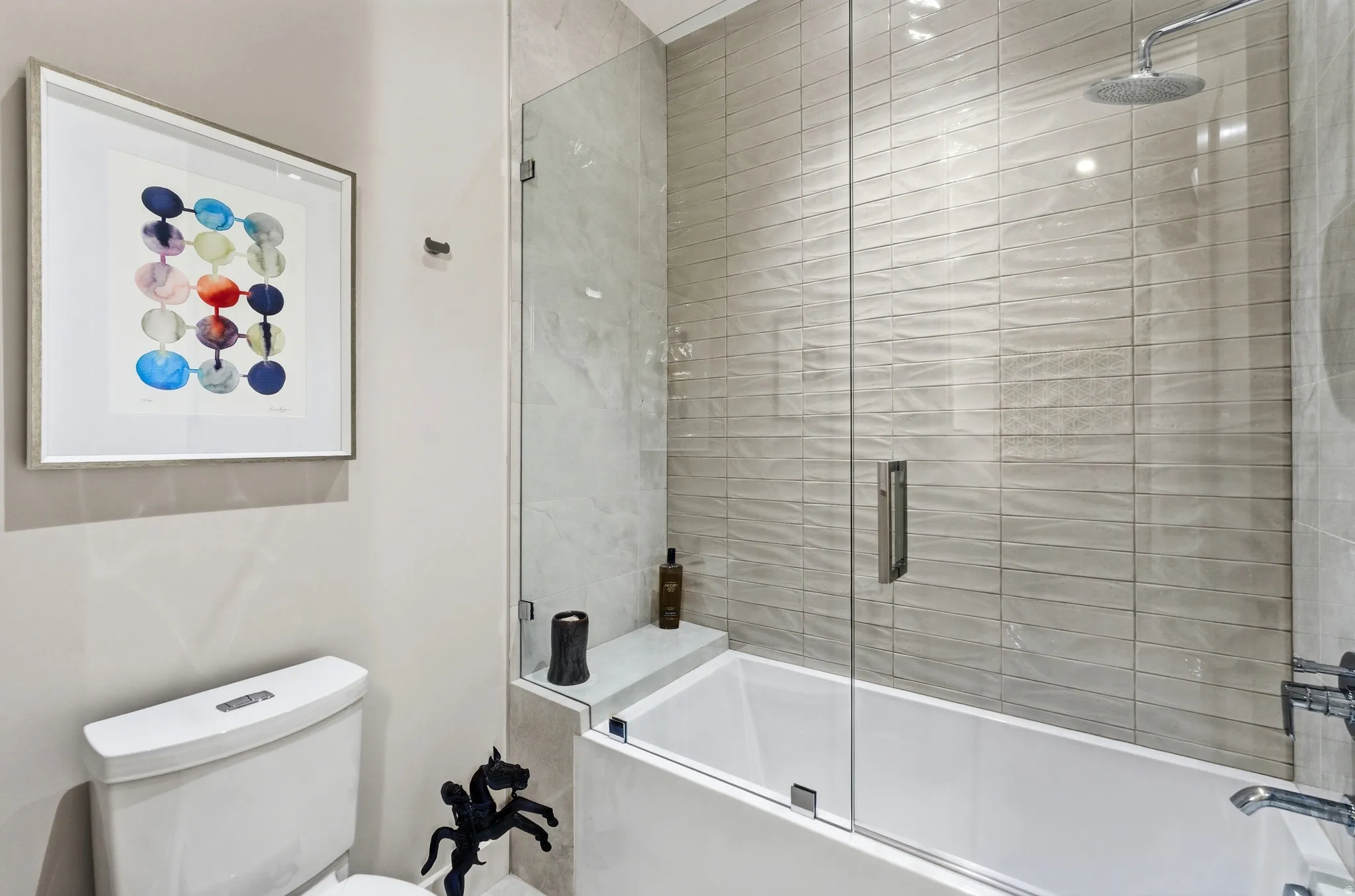 Contemporary bathroom featuring a white porcelain toilet, a tub with a glass enclosure, textured subway tile walls, a rain shower head, and a built-in tub shelf