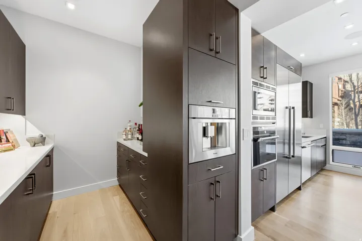 Modern kitchen with warm wood-finish flooring, dark wood cabinetry, white countertops, built-in stainless steel appliances, and an integrated coffee system