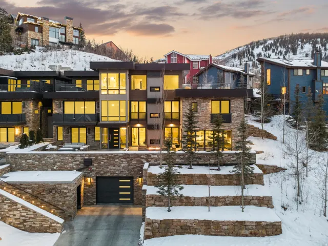 Multi-level contemporary residence featuring extensive stone veneer, expansive window walls, multiple balconies with glass railings, a recessed garage with a modern door, and illuminated exterior steps