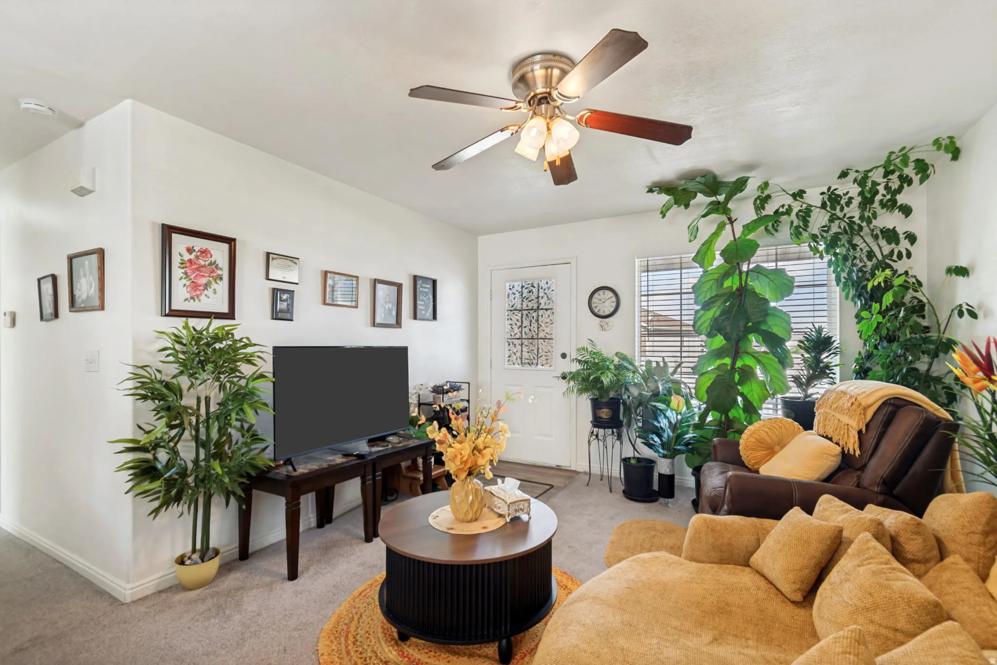 Living area featuring light-toned carpeting, white walls, a ceiling fan with light fixture, and a front entry door with decorative glass