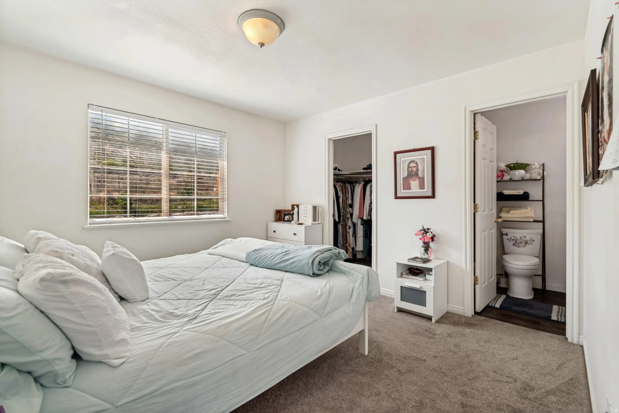 White bedroom featuring a window with blinds, a closet with bi-fold doors, and an attached bathroom with a toilet