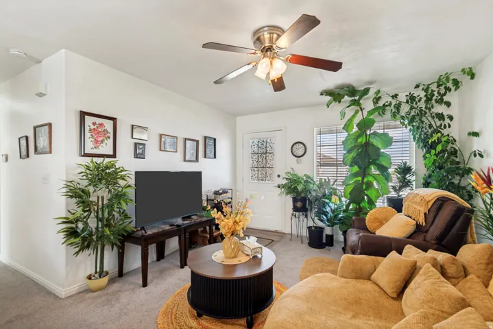 Living area featuring light-toned carpeting, white walls, a ceiling fan with light fixture, and a front entry door with decorative glass