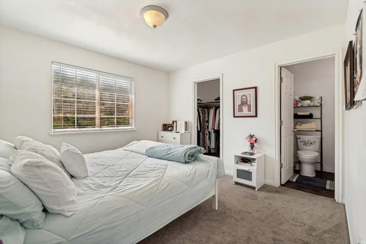 White bedroom featuring a window with blinds, a closet with bi-fold doors, and an attached bathroom with a toilet