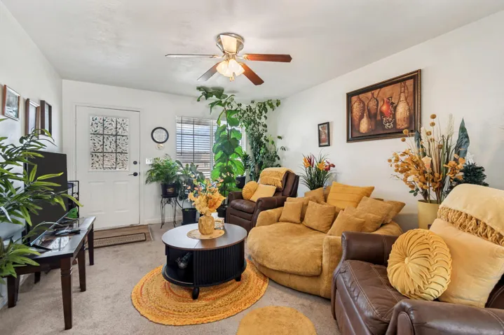 Carpeted living area featuring a white entry door with decorative glass inserts, a window with horizontal blinds, a ceiling fan with integrated lighting, and a dark wood-finish coffee table