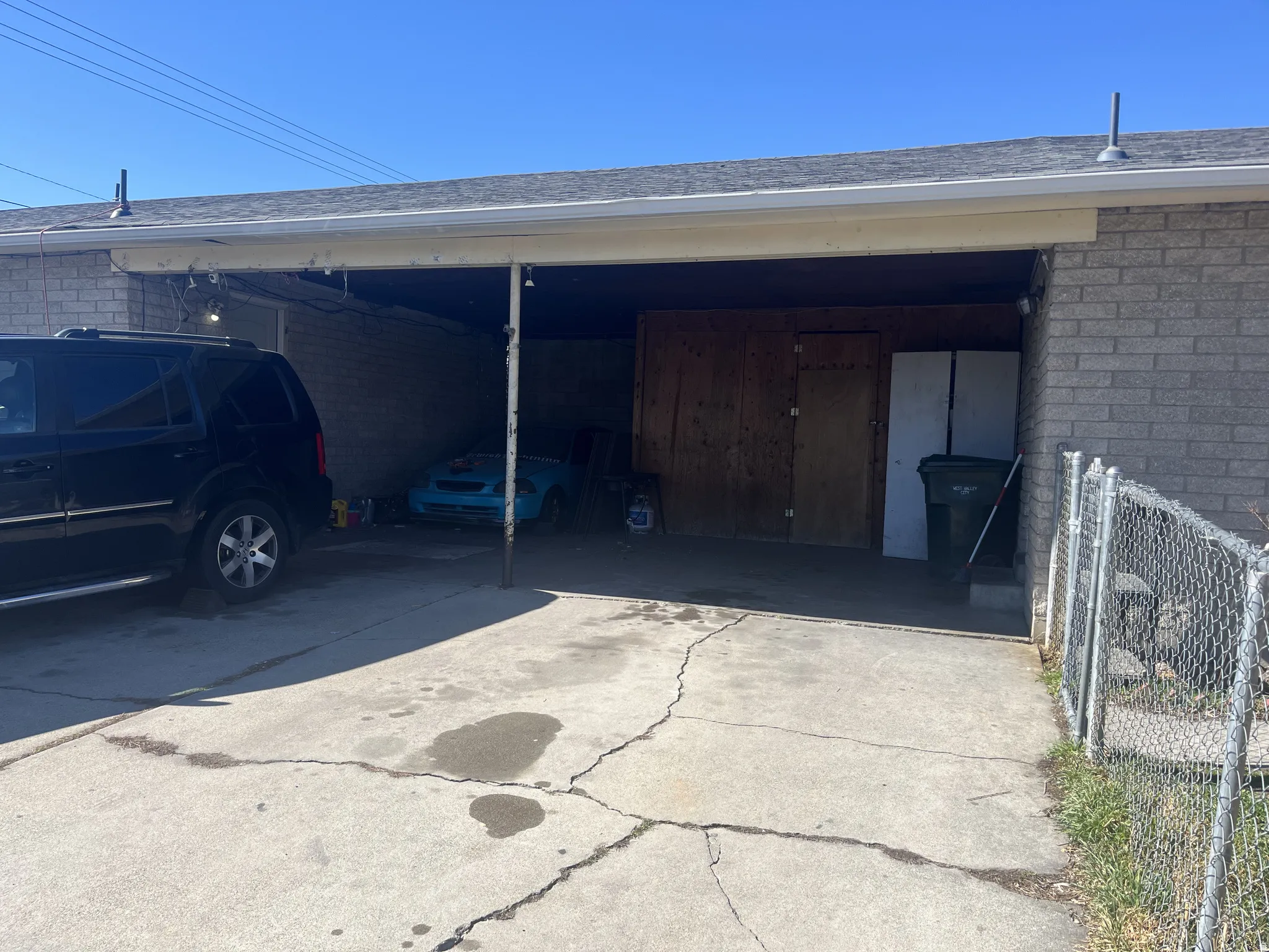 Covered carport area featuring a concrete drive, a chain-link fence, and a gray brick exterior wall