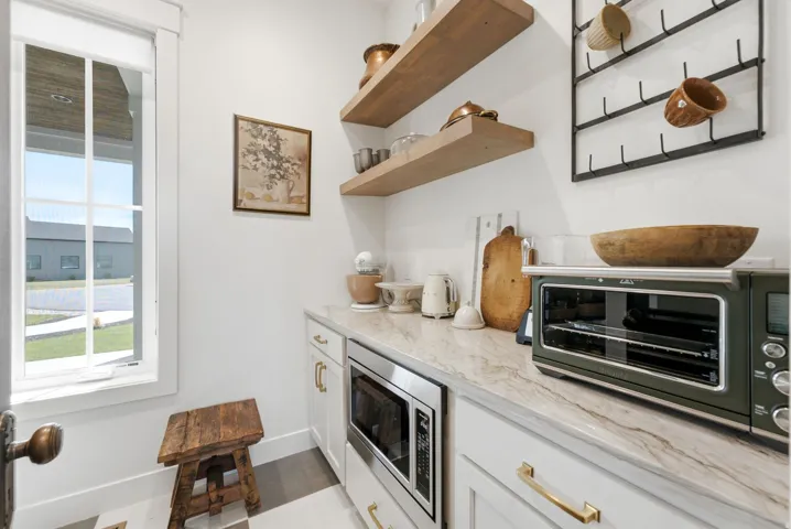 Built-in microwave, stone countertop, white cabinetry with brass hardware, open wood shelving, and a multi-pane window