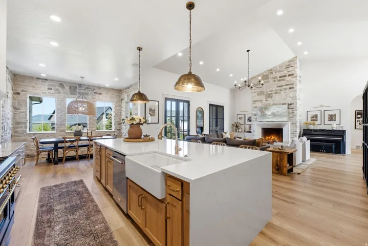 Spacious open-concept living area featuring an expansive kitchen island with a farmhouse sink, wood-finish flooring, a stone-accented fireplace, and vaulted ceilings