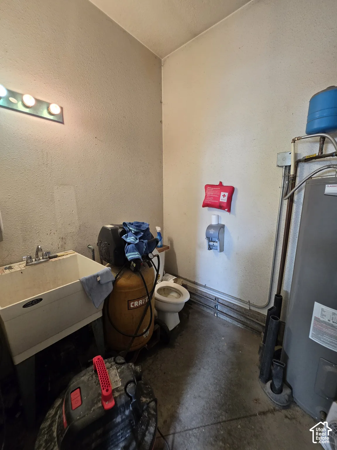 Utility area featuring a deep-basin laundry sink with dual-handle faucet, wall-mounted multi-bulb light fixture, porcelain toilet, and hot water heater