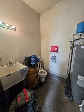 Utility area featuring a deep-basin laundry sink with dual-handle faucet, wall-mounted multi-bulb light fixture, porcelain toilet, and hot water heater