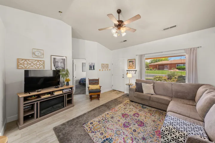 Vaulted ceiling living area featuring a ceiling fan, a large window with blinds, and wood-finish flooring