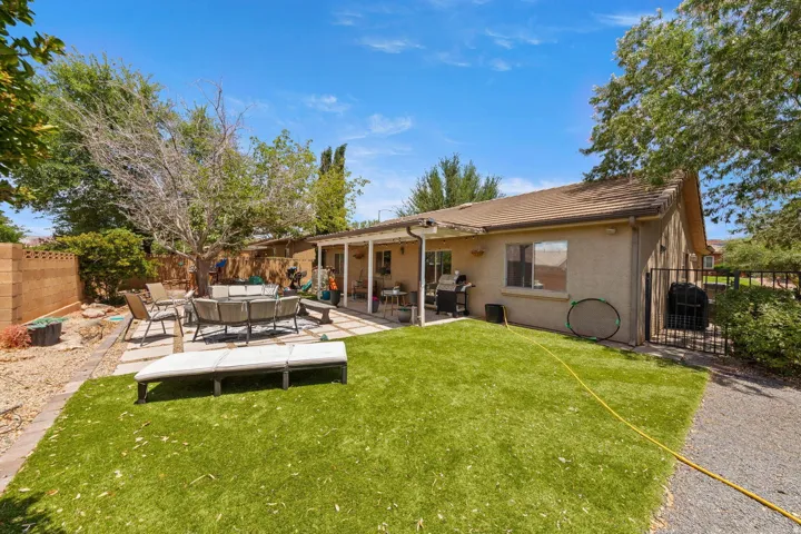 Expansive backyard featuring artificial turf, a large paver patio, a covered patio area, mature trees, and a wrought iron gate