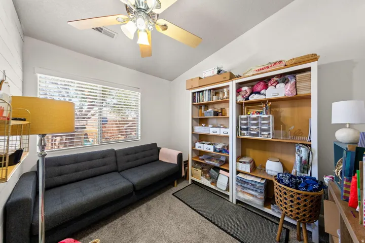 Room featuring a vaulted ceiling with a ceiling fan, a large window with horizontal blinds, built-in shelving, and carpet flooring