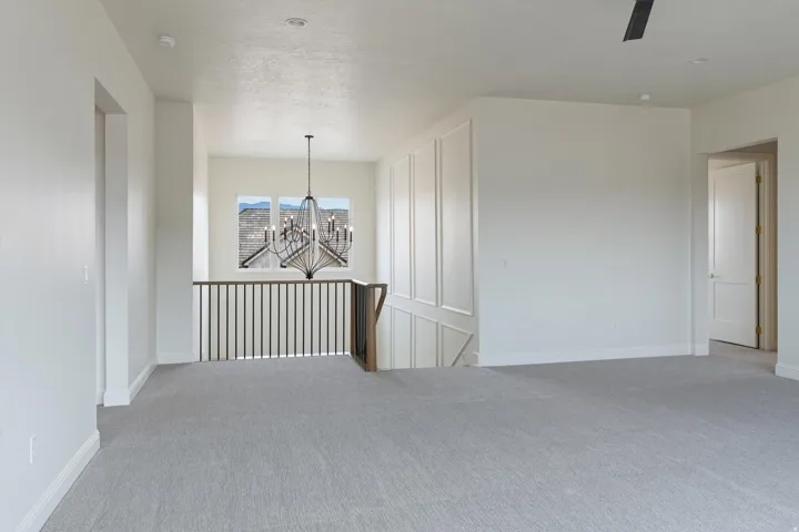 Spacious upper-level landing featuring light gray carpeting, crisp white walls, a decorative chandelier, and a dark wood and metal baluster railing