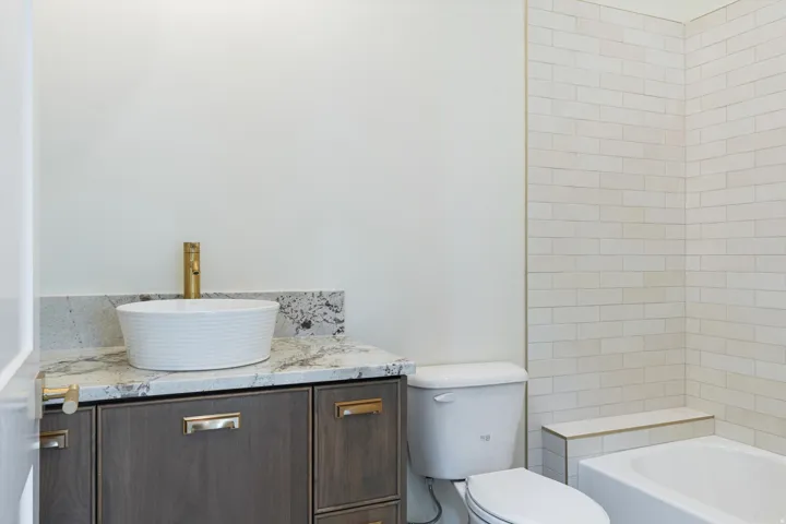 Bathroom featuring a dark wood-finish vanity with a stone countertop, a vessel sink with a gold-tone faucet, a white toilet, and subway tile surrounding the tub
