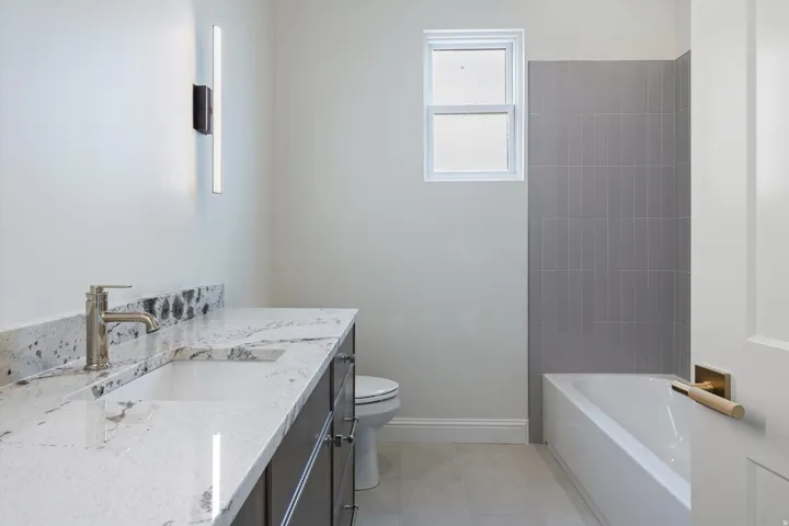 Bathroom featuring a vanity with a white countertop and undermount sink, a sleek wall-mounted light fixture, a white window, a bathtub with grey subway tile surround, and light-toned flooring