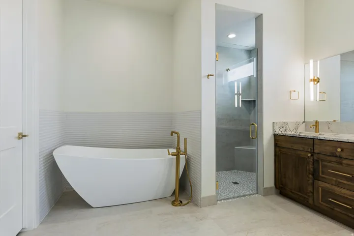 Freestanding soaking tub with floor-mounted faucet, walk-in shower with glass enclosure, custom wood-finish vanity with stone countertop, and dual vertical wall sconces