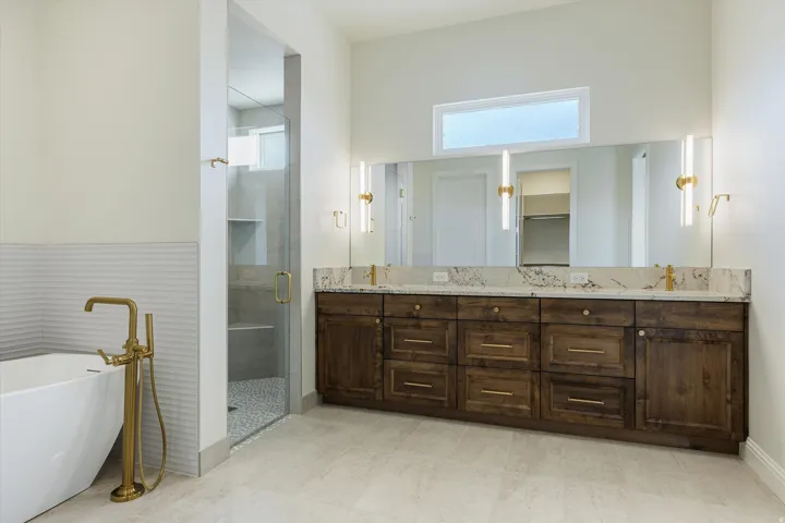 Freestanding soaking tub with a floor-mounted brass faucet, glass-enclosed shower, dual vanity with a stone countertop, extensive wood cabinetry, and tiled flooring