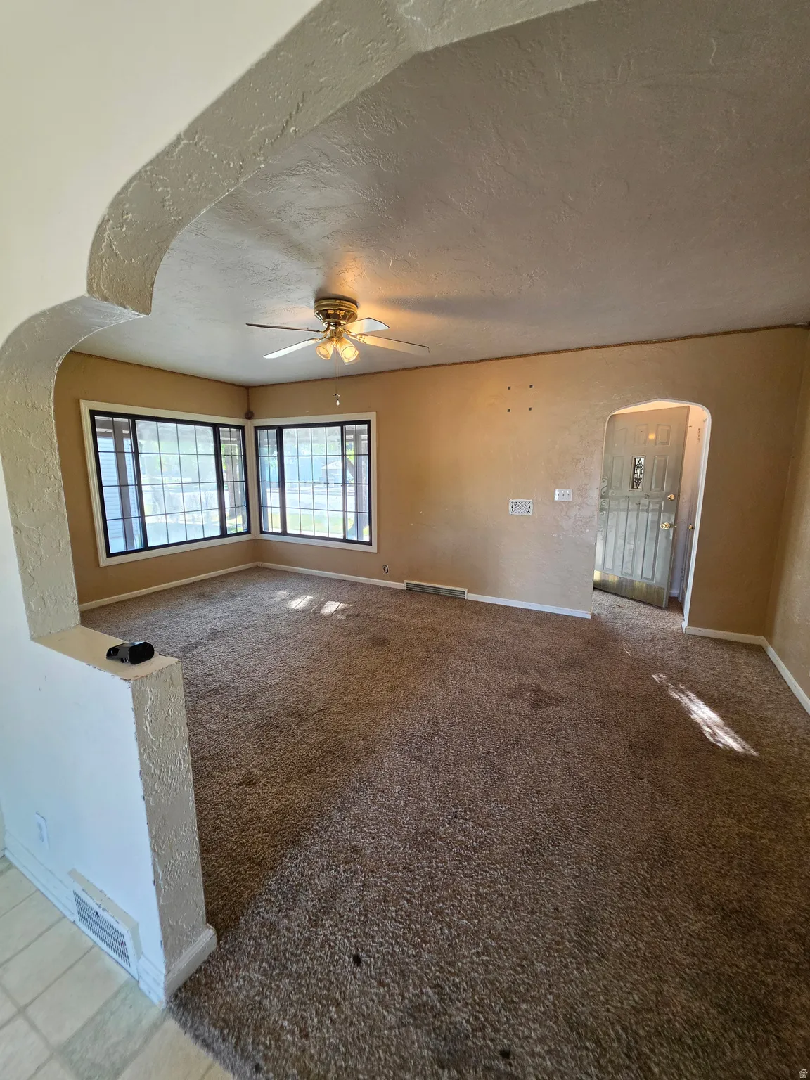 Arched entryway leading to a carpeted room with multiple windows, a ceiling fan, and a light-colored wall finish