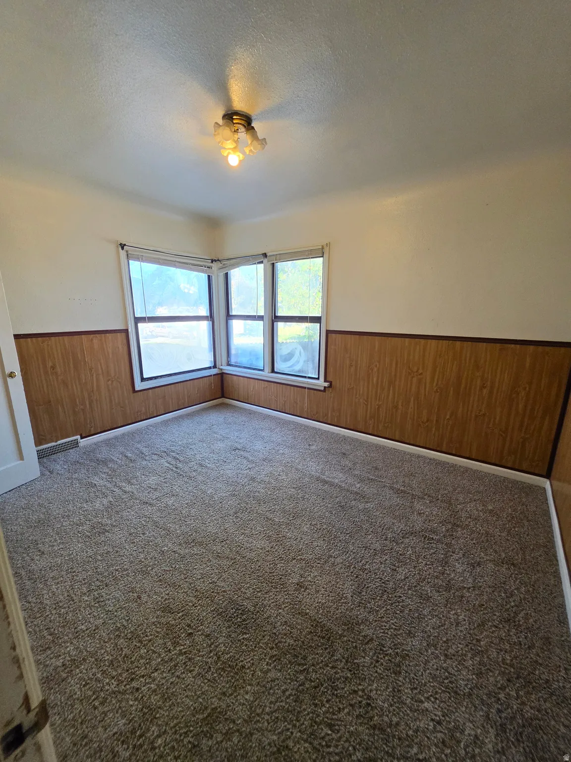 Corner windows providing ample natural light, wall paneling, ceiling-mounted light fixture, and carpeted flooring