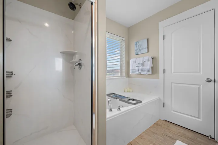 Spacious bathroom featuring a glass-enclosed shower with white marble-look paneling, an integrated soaking tub with matching marble-look surround, a window with blinds, and wood-finish flooring