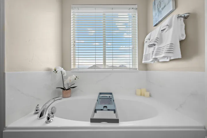 Corner soaking tub featuring polished chrome fixtures, white marbled-effect tile surround, and a window with horizontal blinds