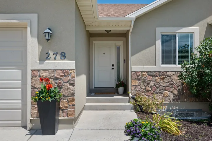 Stucco and stone facade featuring a white entry door with a sidelight, a garage door, a window with blinds, and established landscaping