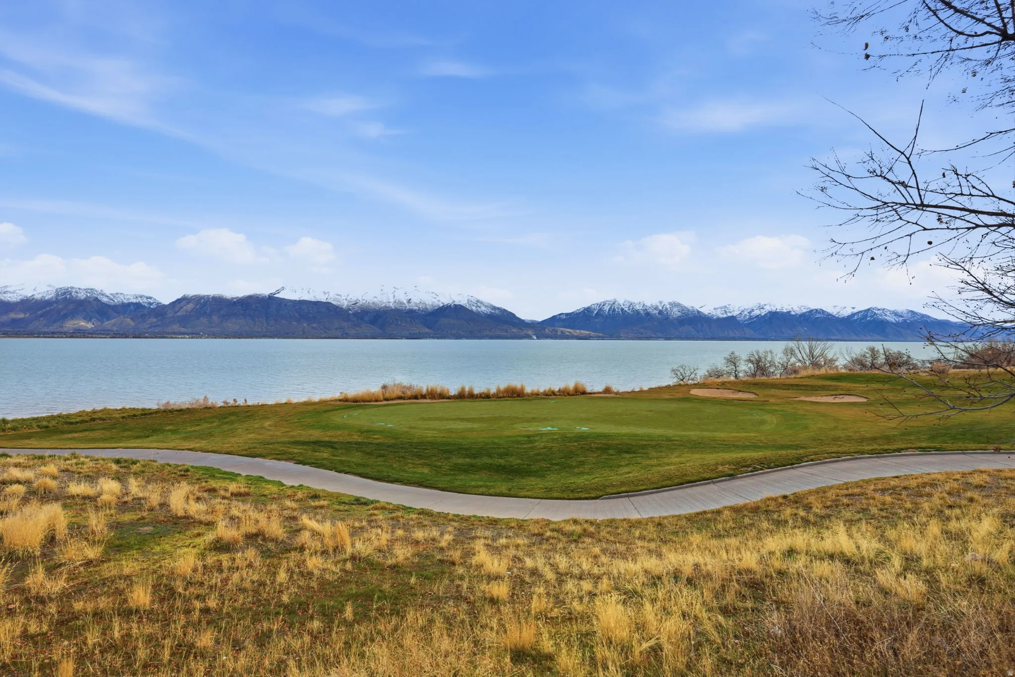 Expansive golf course green with sand traps bordering a large body of water, set against a backdrop of snow-capped mountains under a clear blue sky