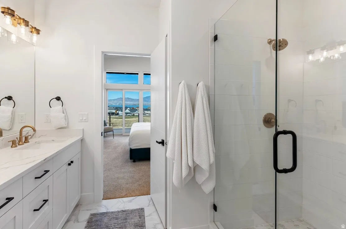 Bathroom featuring a spacious walk-in shower with clear glass enclosure and bronze fixtures, a vanity with a light-toned countertop and gold-toned faucet, and crisp white cabinetry with black hardware