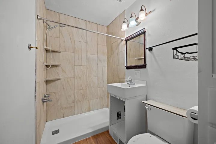 Bathroom featuring a tiled shower surround, built-in shower shelving, vanity with a rectangular sink, wall-mounted mirror, and wood-finish flooring