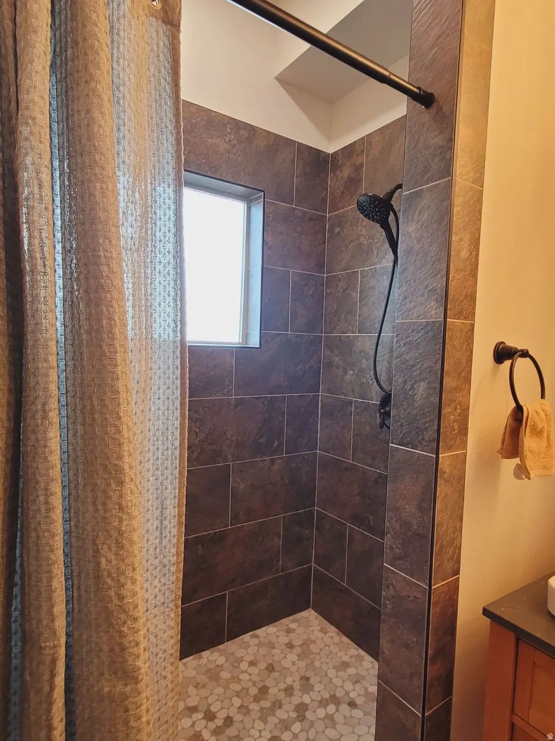 Shower stall featuring large format wall tiling, pebble-style floor tiling, matte black shower fixtures, and a recessed window