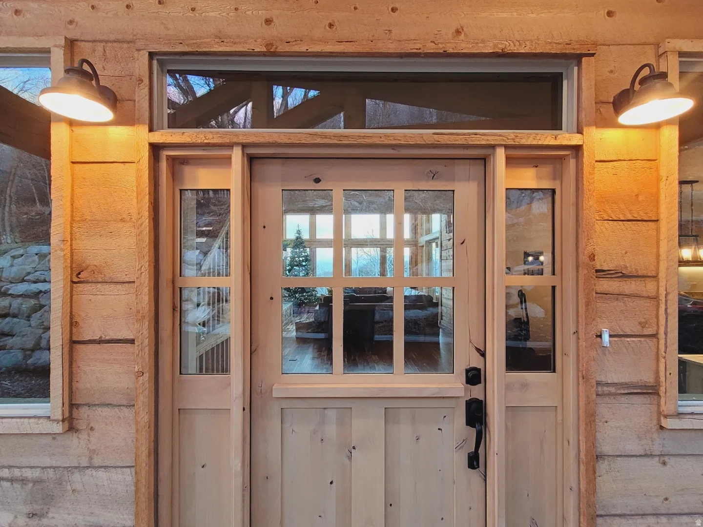 Crafted wood-paneled door with multiple window panes and sidelights, complemented by a transom window and rustic wall sconces