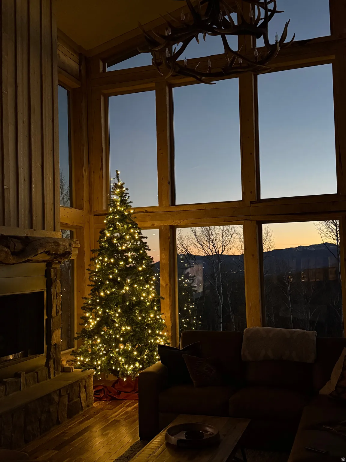 Expansive window wall with mountain scenery, featuring a prominent antler chandelier, wood-finish flooring, and a stone-clad fireplace with a television inset