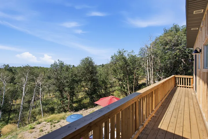 Spacious outdoor deck featuring natural wood flooring and railing, with exterior wall lighting, overlooking a natural treed landscape