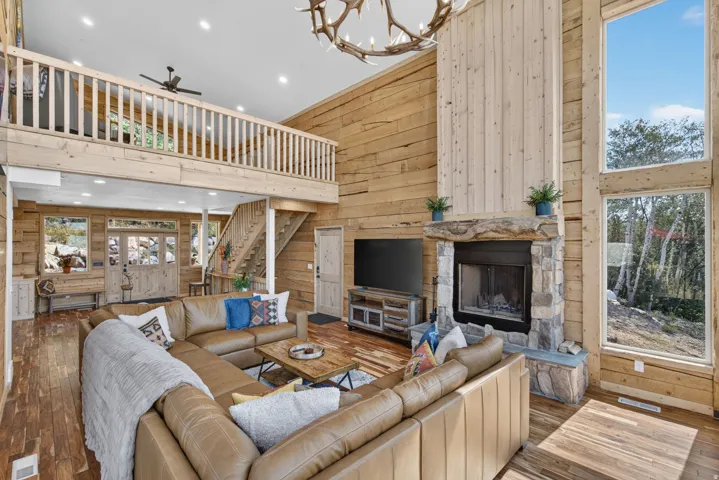 Soaring wood-paneled great room featuring a two-story stone fireplace with integrated media console, leather sectional seating, and expansive windows overlooking forested landscape
