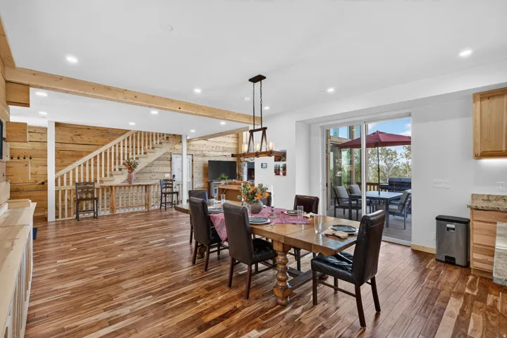 Spacious dining area featuring wood-finish flooring, a contemporary chandelier, recessed lighting, a sliding glass door to an outdoor deck, and a wood accent wall with a staircase