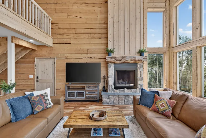 Open concept living space featuring a two-story stone fireplace with a wood mantle, expansive wood-frame windows, wood-finish flooring, and a second-level open-rail balcony