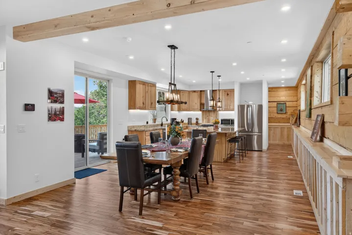 Open-concept dining and kitchen area featuring wood-finish flooring throughout, custom wood cabinetry with stainless steel appliances, a wood dining table with seating for six, pendant lighting, exposed wood beam ceiling detail, recessed lighting, and dir
